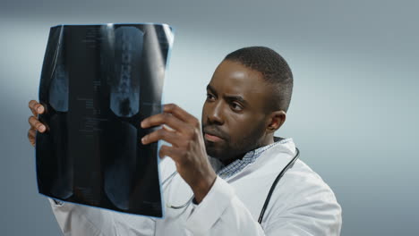 Close-up-of-the-young-African-American-man-doctor-or-intern-holding-X-ray-copy-and-studying-it-carefully-on-the-grey-background.